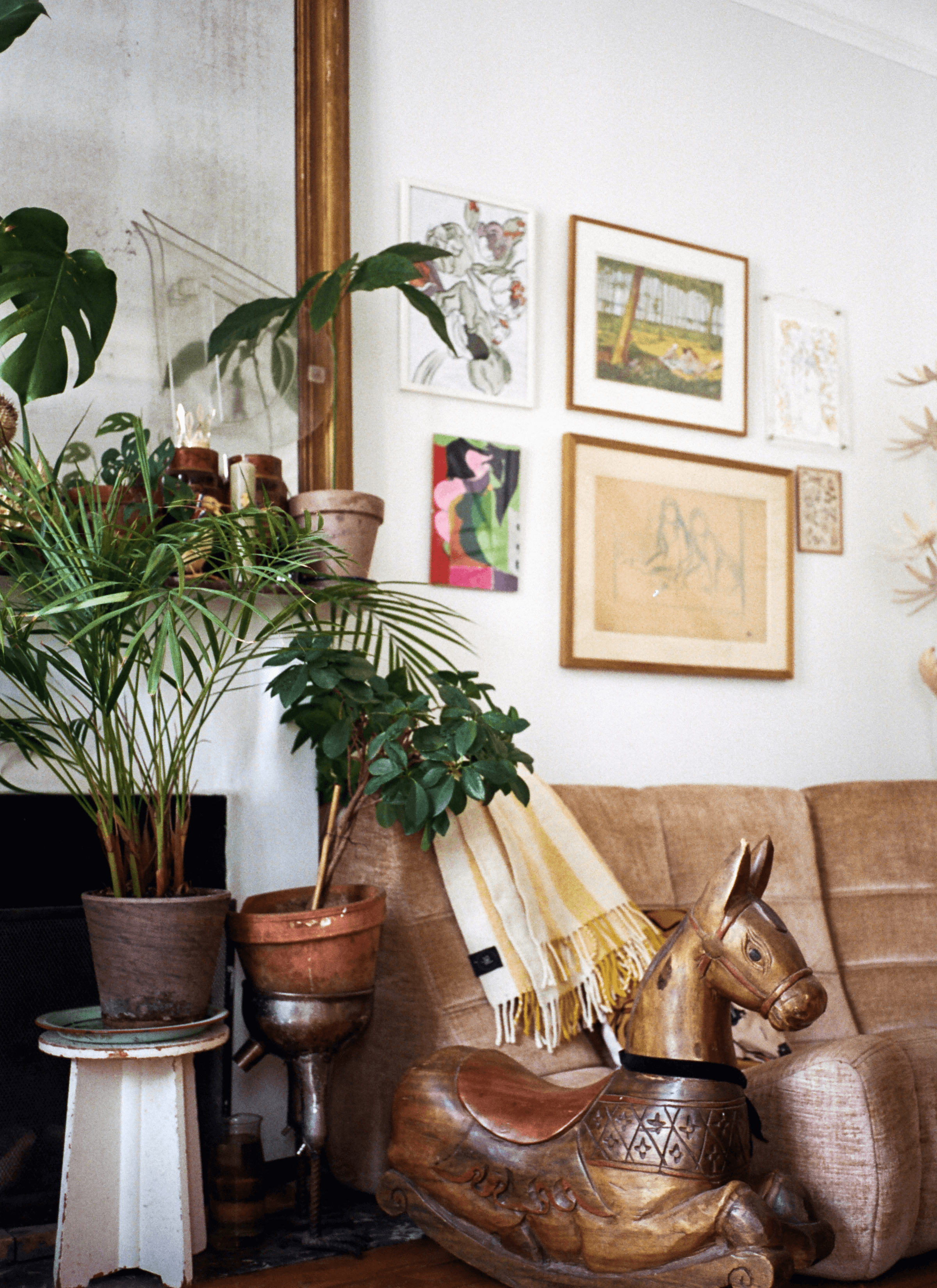 A collection of potted plants decorated the room next to a sofa with a wooden-like horse statue. There are some art and framed pictures being hung on the wall.