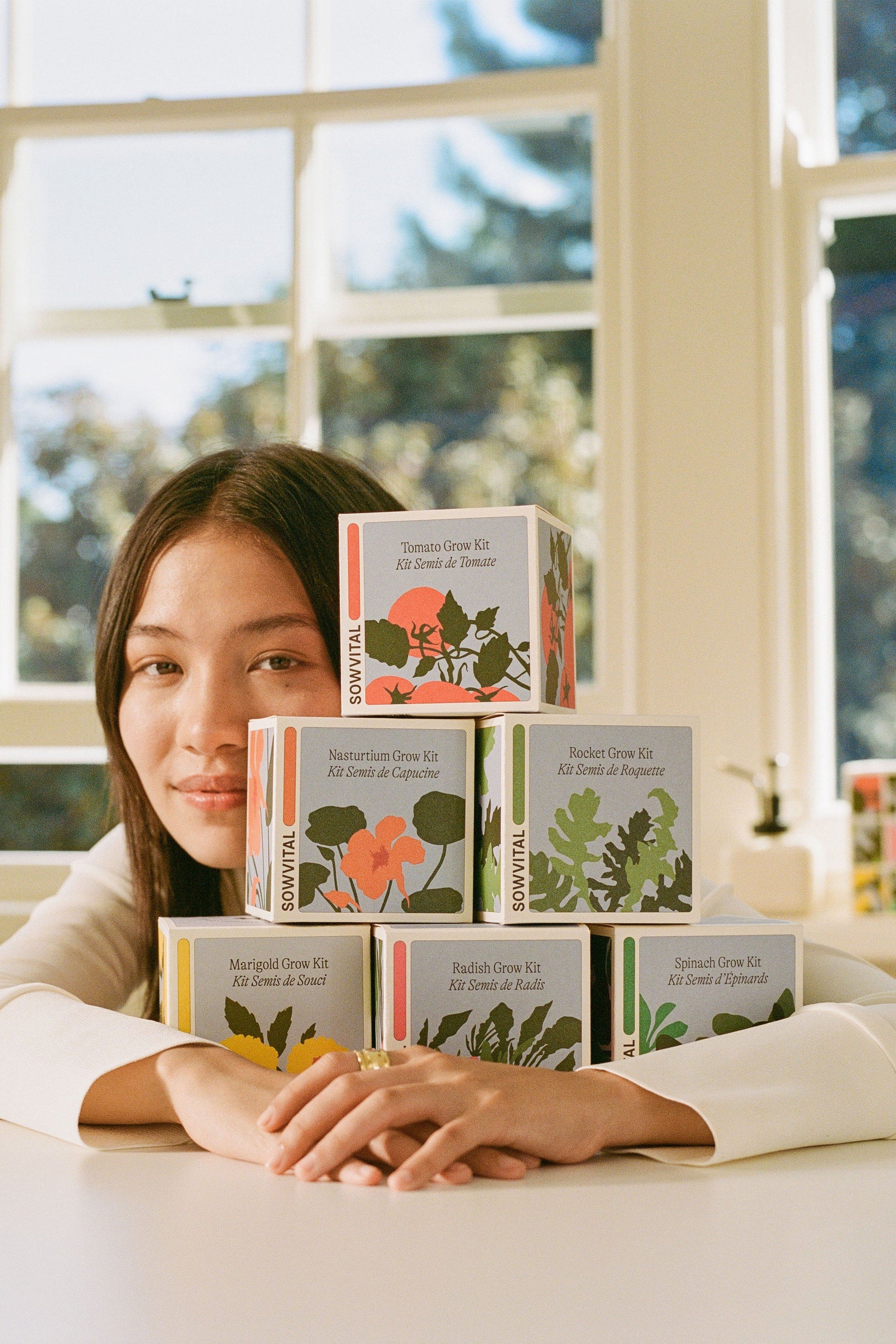 A stack of Sowvital Seed Grow Kits with a female model wrapping her arms around them from behind, her face peeking from behind the stack. There are 6 varieties of grow kit, Tomato, Nasturtium, Rocket, Marigold, Radish, and Spinach. 