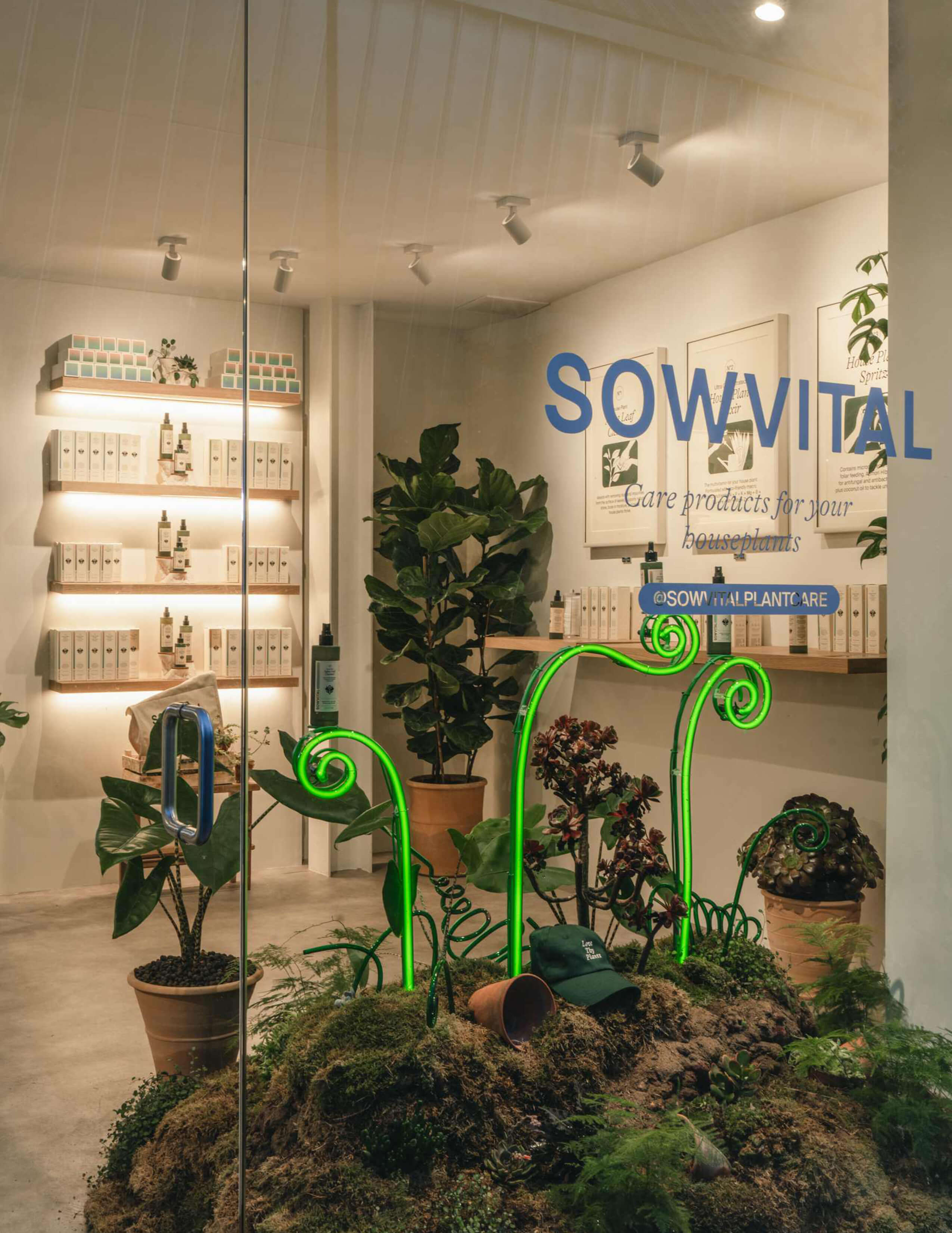 A front side angle of the pop-up store with the Sowvital logo printed at the front and a big pile of soil decorated with different kinds of plants and green LED light. The products are presented on the shelves to see through from the glass.