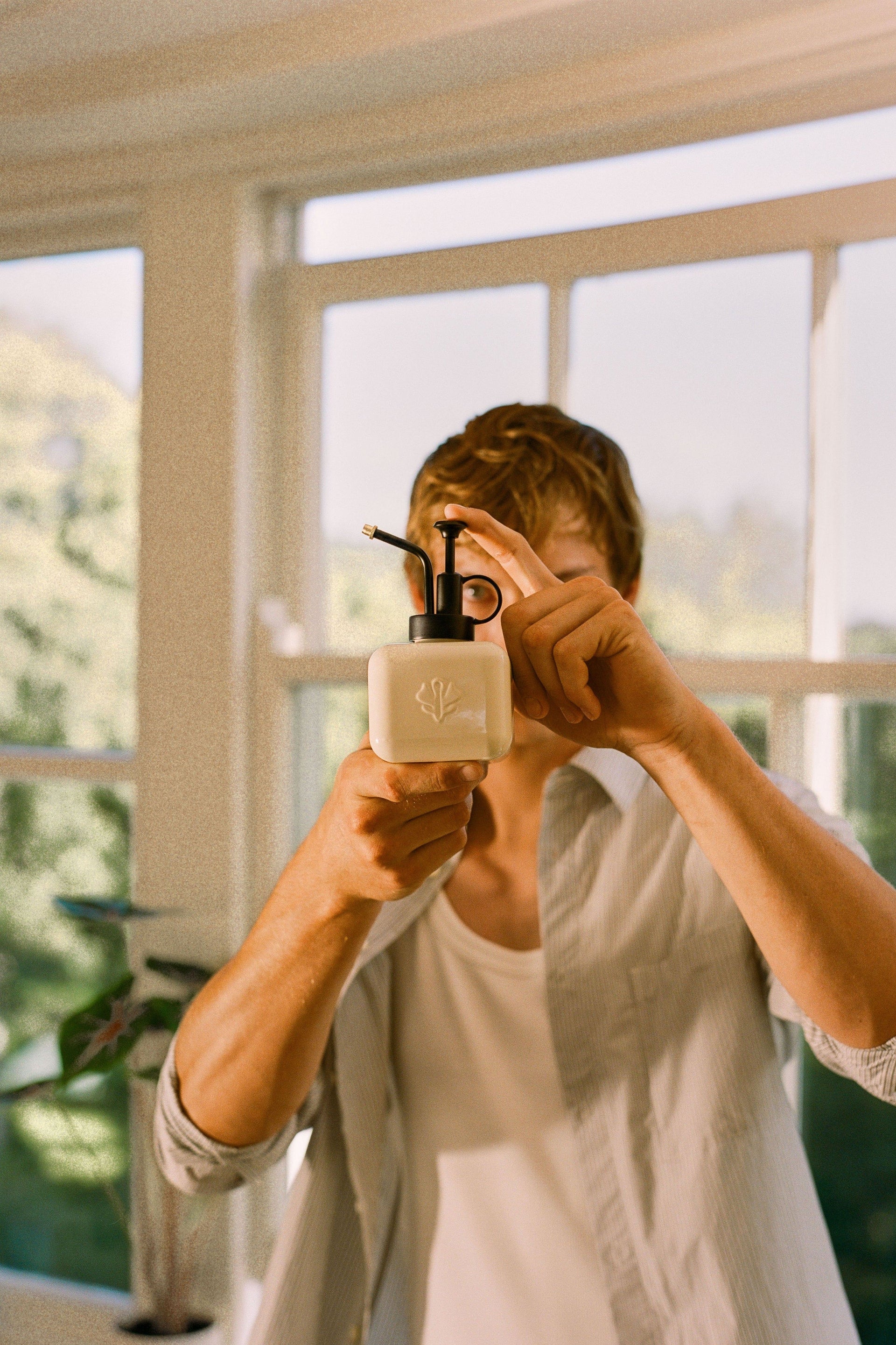 A male model holds a beige stainless steel houseplant mister up, looking through the handle like a camera.