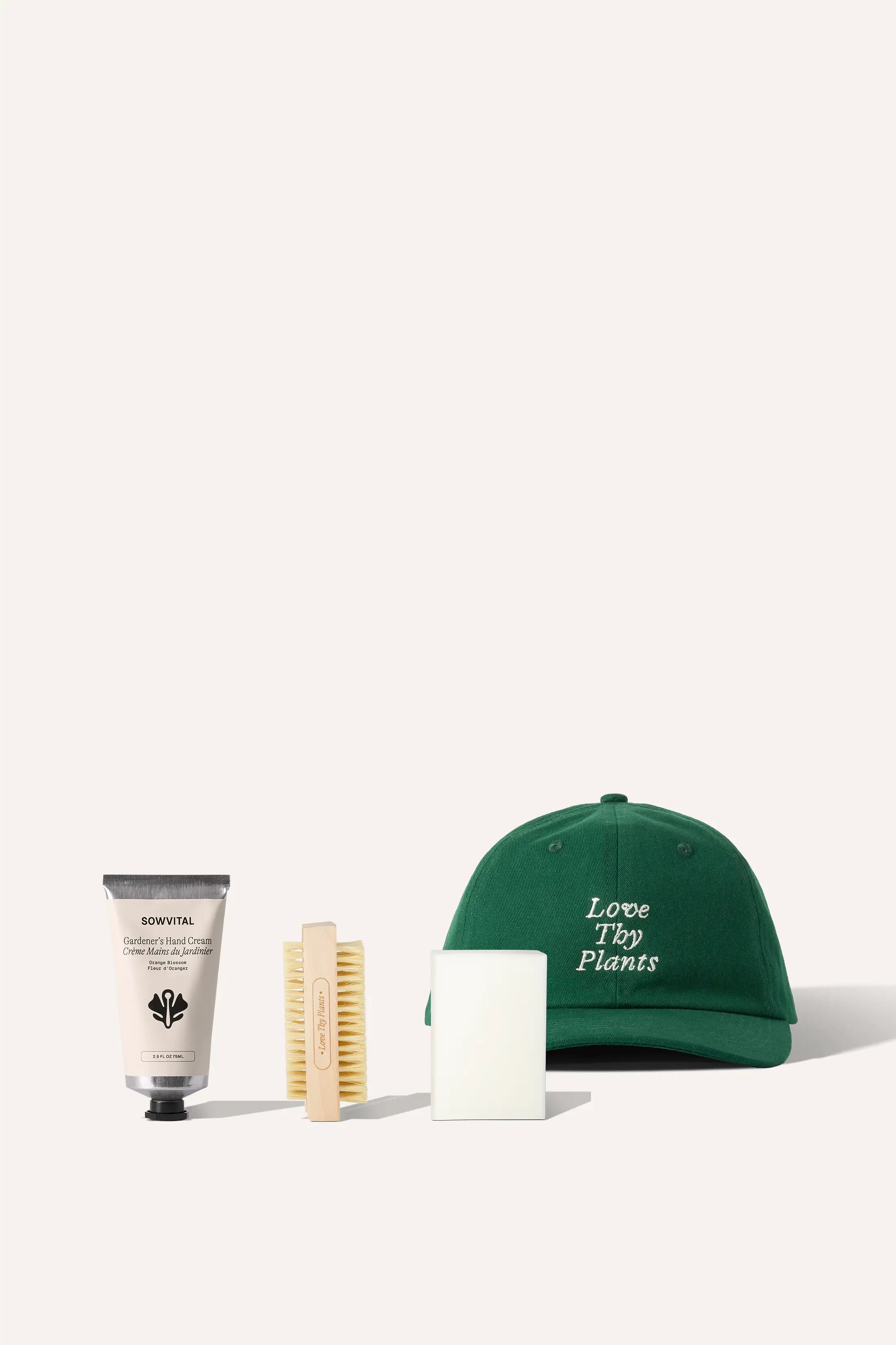 A tube of hand cream, nail brush, and bar of soap sit in front of a green cap embroidered with the phrase "Love Thy Plants".