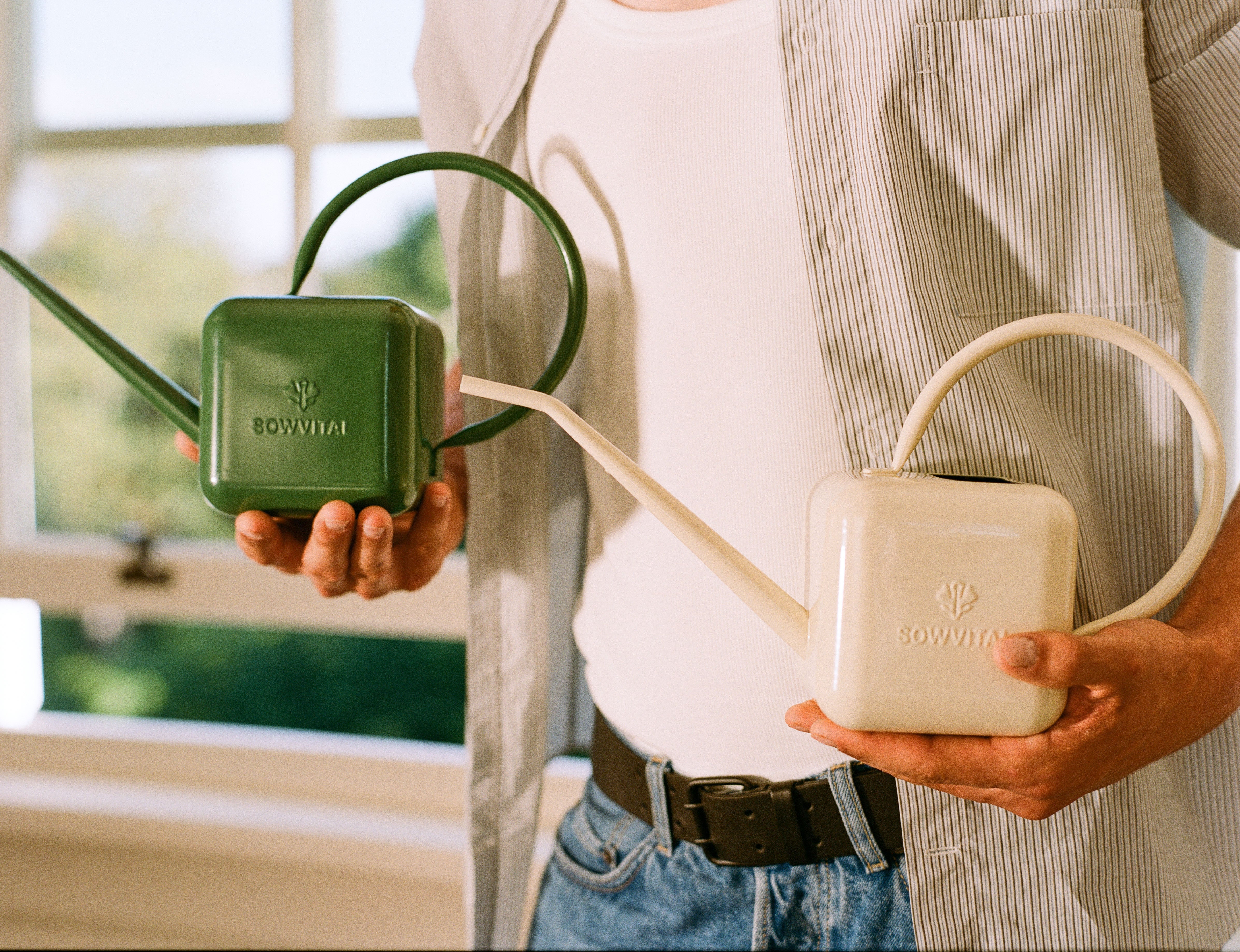 A male model holds two Sowvital watering cans at his waist. One is beige and one is green. They are made of stainless steel.