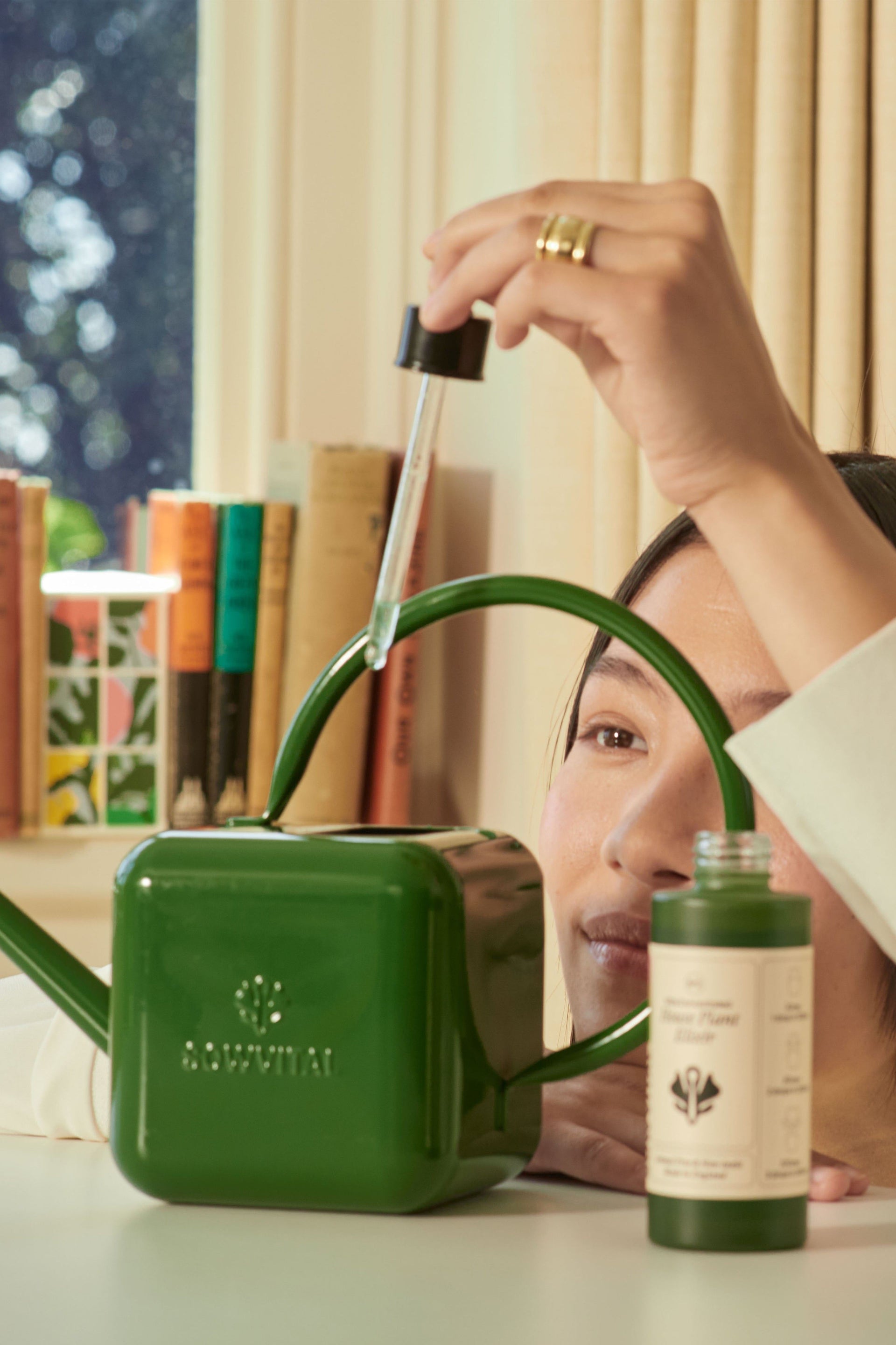 A female model holds the pipette dropper lid of the House Plant Elixir high above a Sowvital stainless steel green watering can, watching as she drops the fertiliser into the watering can.