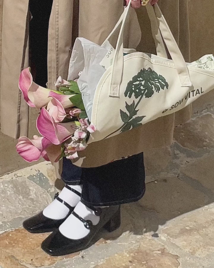 A person wearing a long beige coat holds a cotton flower tote with a bundle of flowers in it, swaying in the wind.